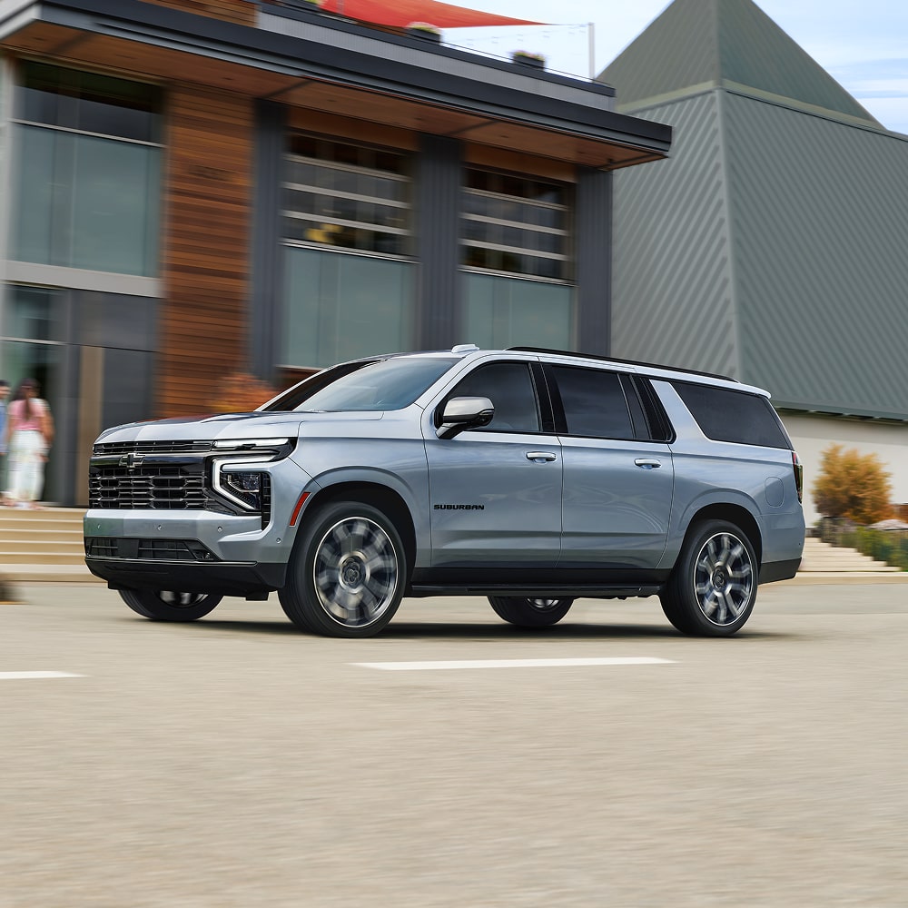 Profile View of the 2026 Chevy Suburban SUV Driving Past a Modern Apartment Building