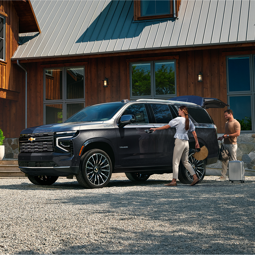 Three-quarters View of a 2026 Chevy Tahoe Parked in a Gravel Driveway with a Family Loading Luggage in the Trunk